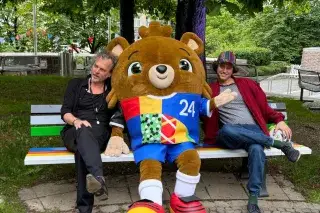 Zwei Männer und EM-Maskottchen Albärt auf der #hockdiher-Bank vor dem Fat Cat München