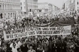 Fotografie einer Menschenmenge, die gegen die bayerische Aids-Politik demonstriert. Zu sehen sind mehrere Banner, unter anderem eines mit der Aufschrift „Wir Schwule schützen uns vor Aids, wer schützt uns vor dieser Aids-Politik?“ Fotografie einer Menschenmenge, die gegen die bayerische Aids-Politik demonstriert. Zu sehen sind mehrere Banner, unter anderem eines mit der Aufschrift „Wir Schwule schützen uns vor Aids, wer schützt uns vor dieser Aids-Politik?“